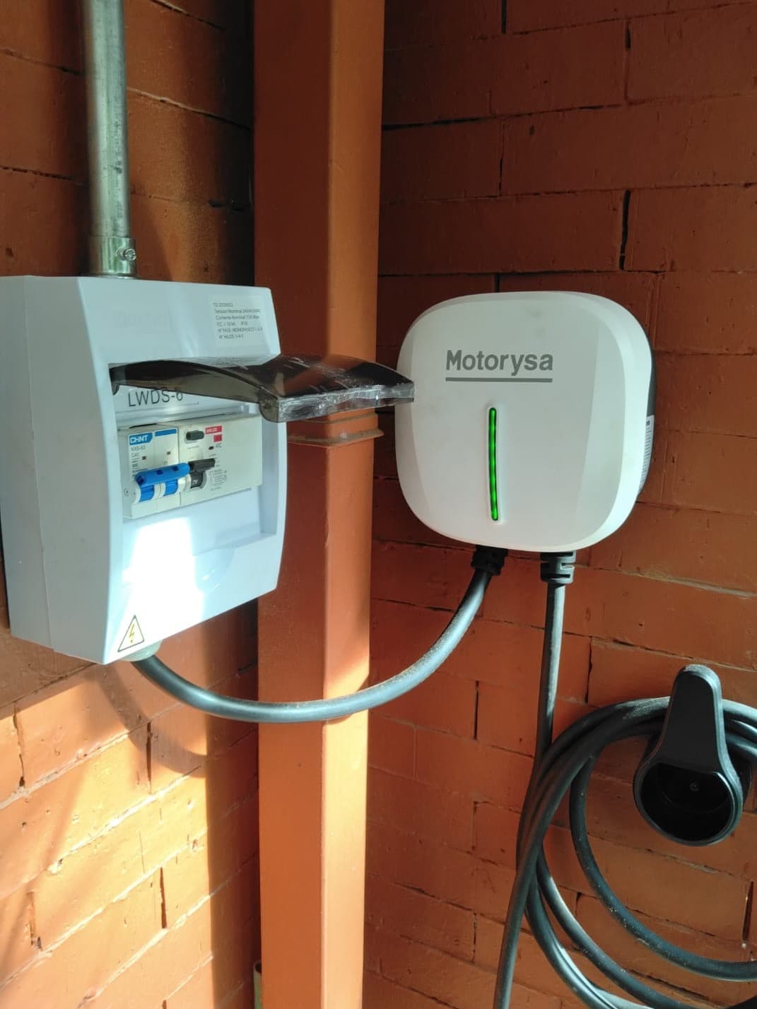 Cargador blanco junto a un tablero con tapa abierta y canalización sobre una pared de ladrillo